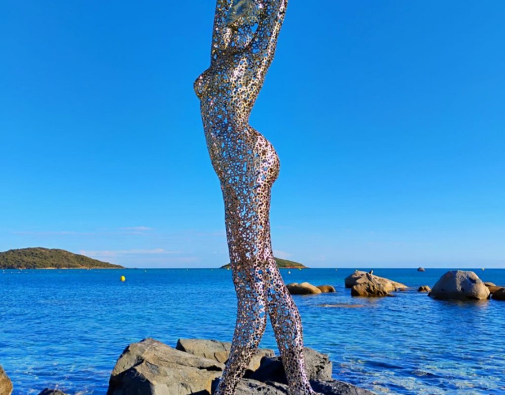 Sculpture en métal d'un corps de femme sur des rochers avec la plage en arrière plan